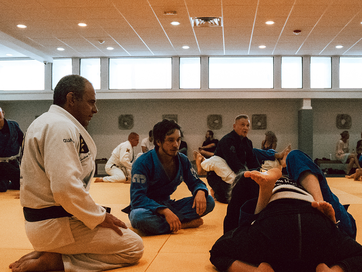 Two Brazilian Jiu-Jitsu students sparring on a mat with two male professors observing in Gustavo Machado’s BJJ Academy.