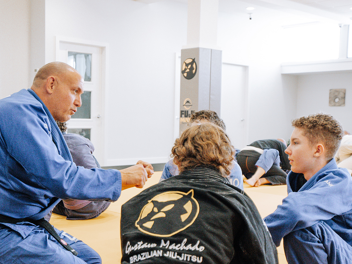 A Brazilian Jiu-Jitsu professor explaining something to a male and female student while sitting down on a mat at Gustavo Machado Academy.