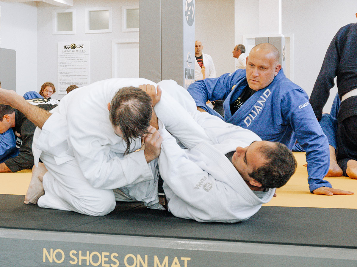 Two Gustavo Machado Academy BJJ students spar on mat with Head Instructor Justin observing behind them.